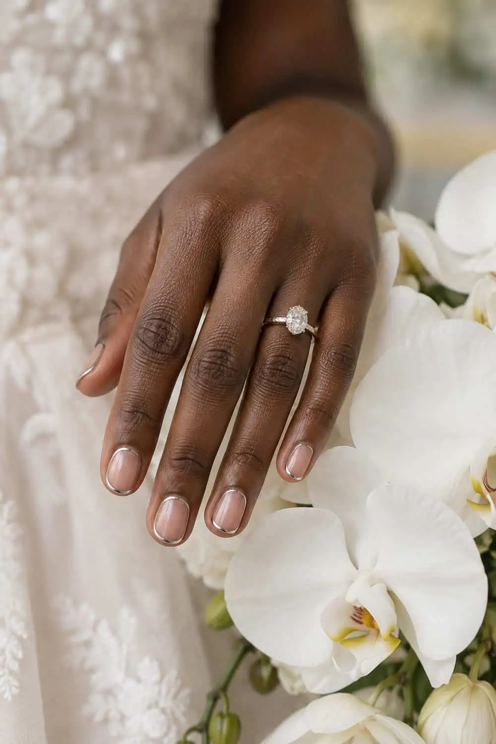 Bridal dark-brown hand with short rounded nails, glossy clear base and very thin silver outline around each edge, beside white orchids
