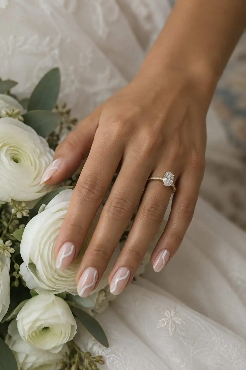 Bridal olive-toned hand with medium oval nails, sheer nude base and thin flowing white swirl lines, beside white ranunculus