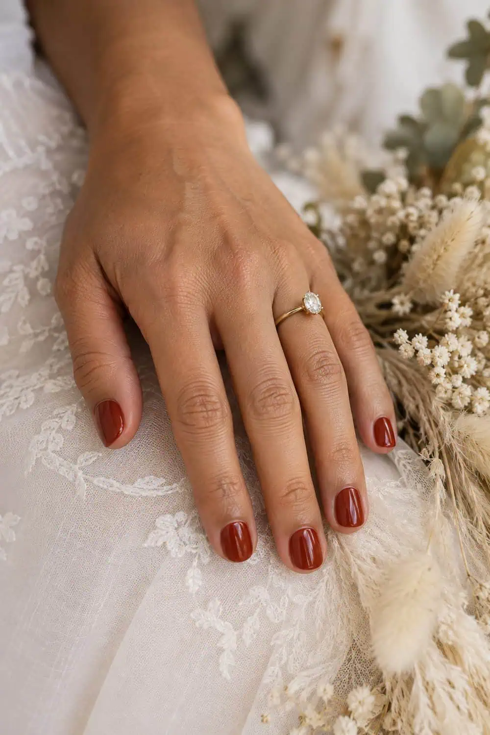 Bridal olive-toned hand with short rounded nails painted solid brick-red polish, dried cream grasses nearby