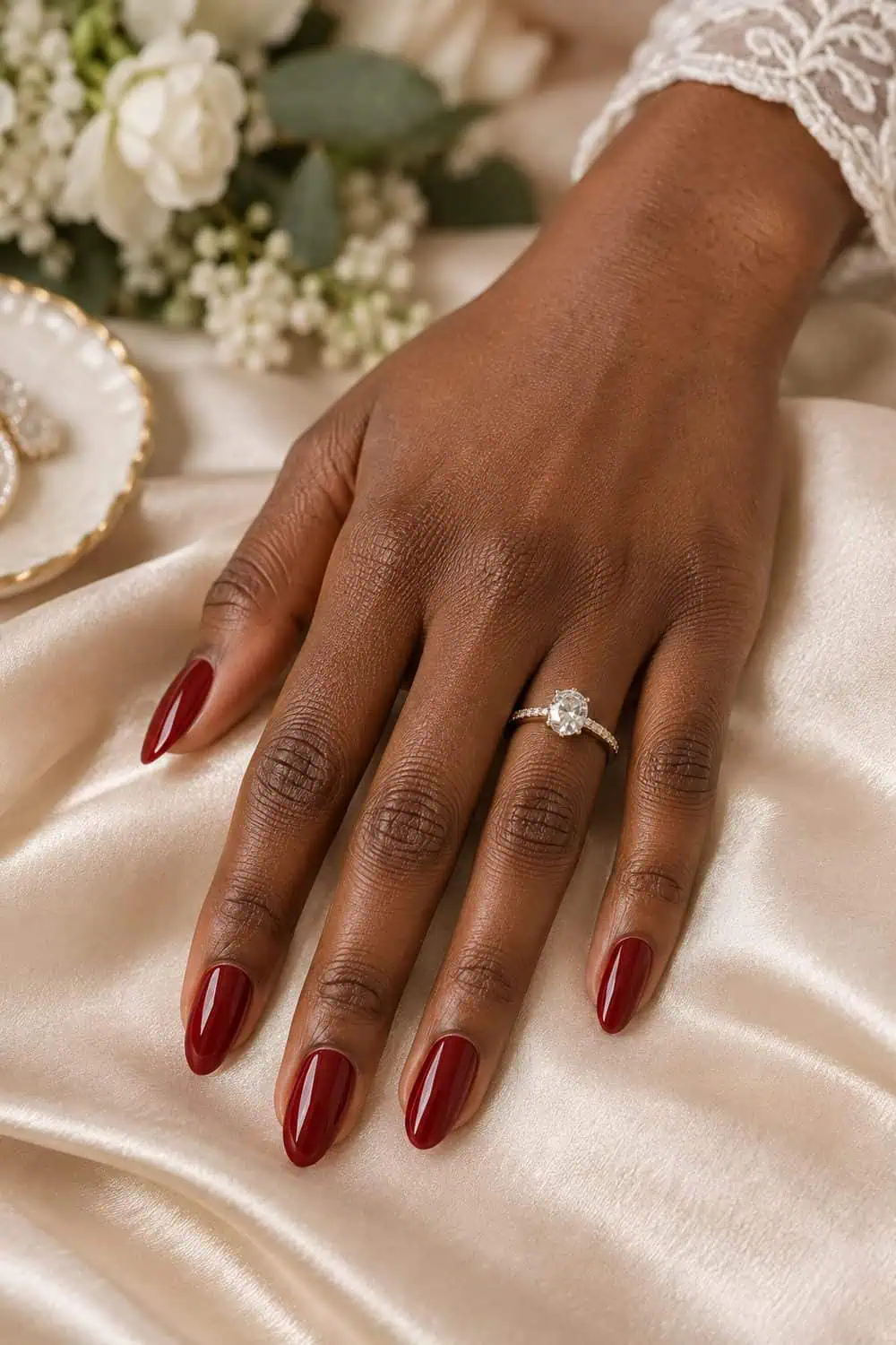 Bridal deep-brown hand with medium almond nails painted solid glossy burgundy-red polish, champagne satin background