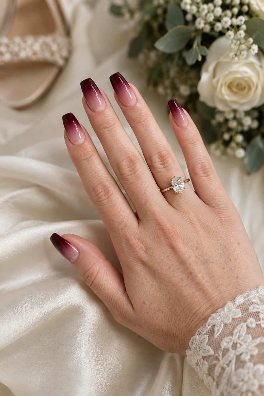 adult bride's fair freckled hand with medium coffin nails fading from deep burgundy tips to sheer pink cuticles, cream satin