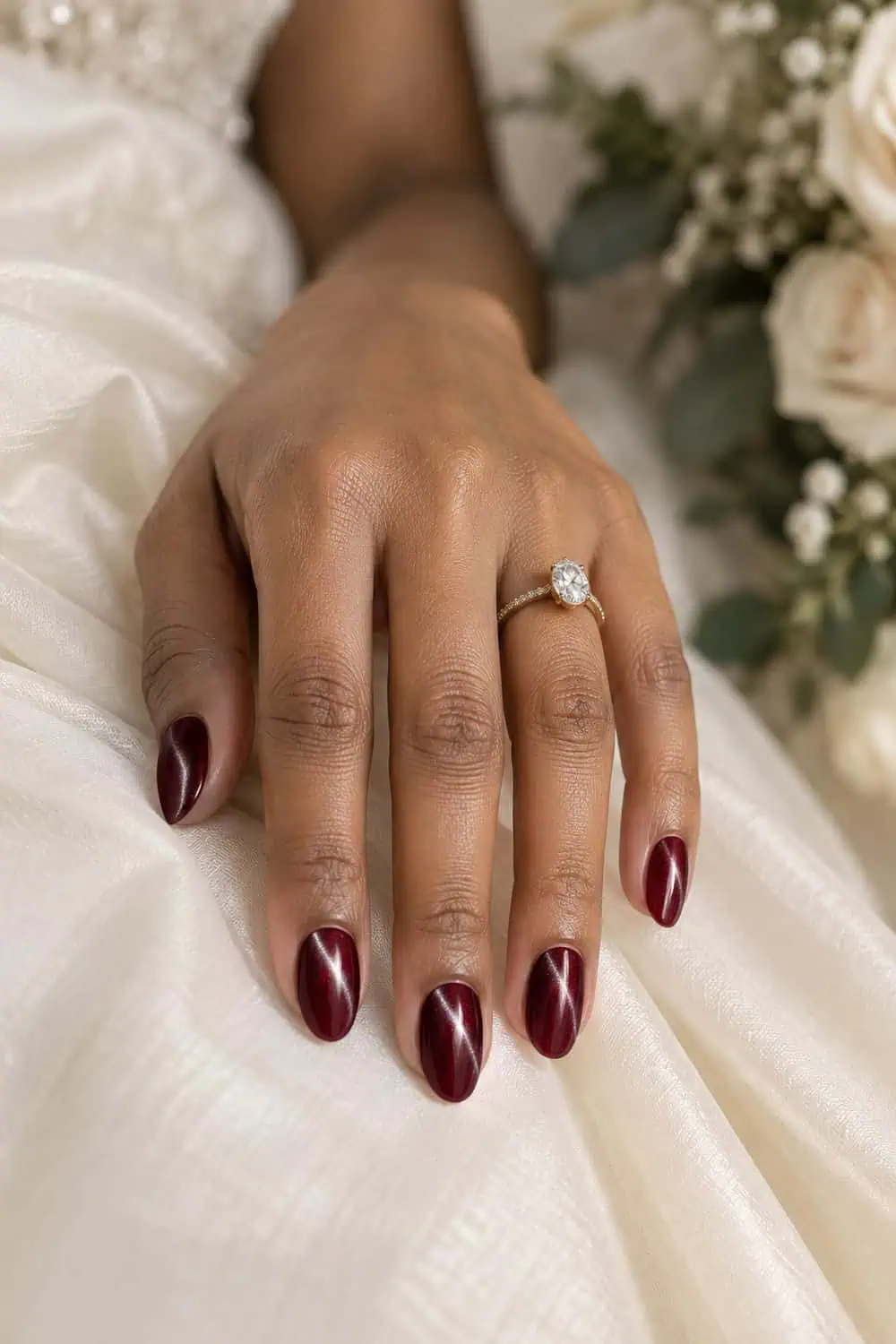 Bridal light-brown hand with short almond nails painted wine-red magnetic velvet polish, narrow light bands visible, ivory fabric