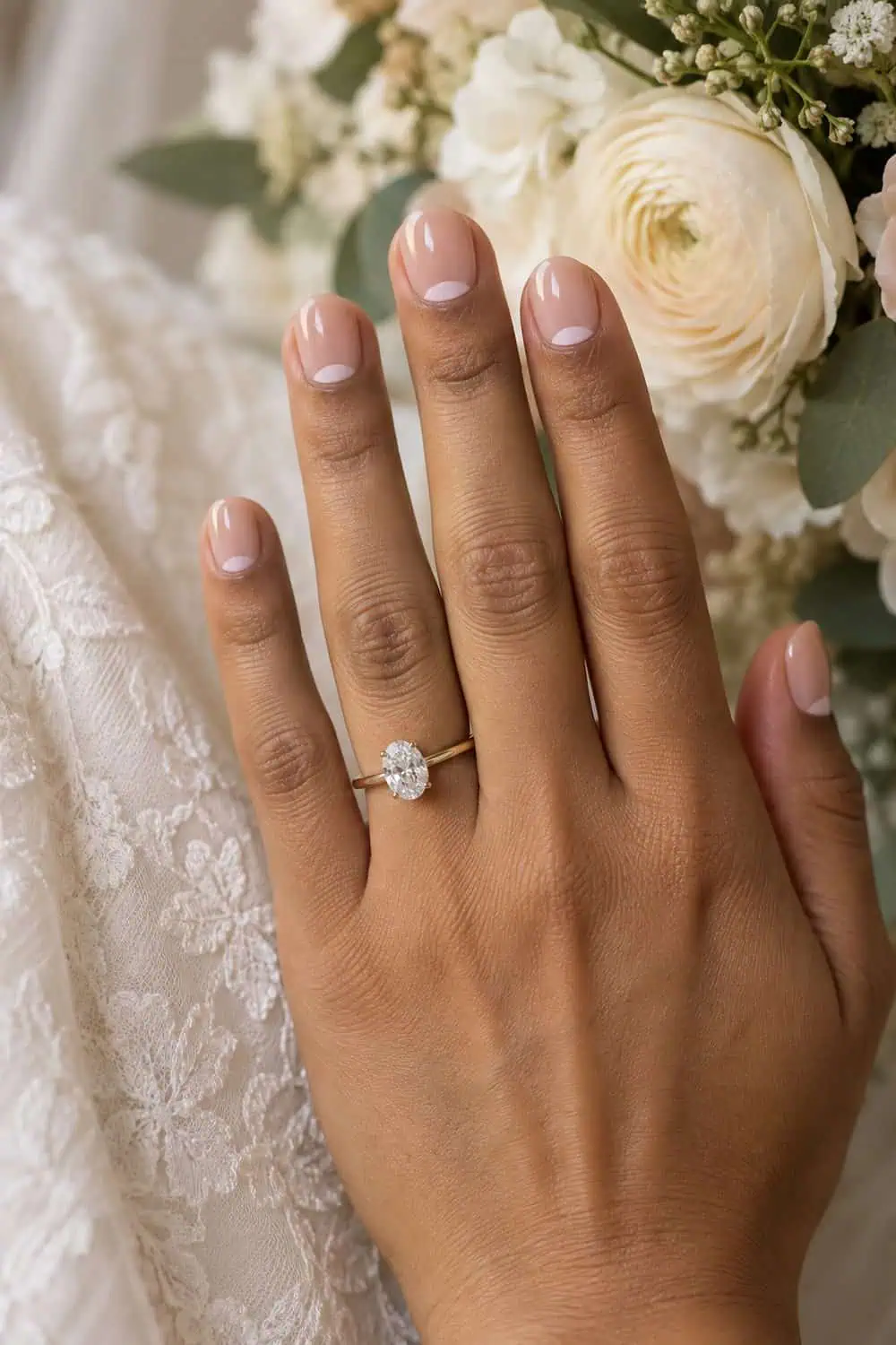 Bridal light-brown hand with short rounded nails, clear gloss and small pale-pink half-moons at cuticles, cream flowers nearby