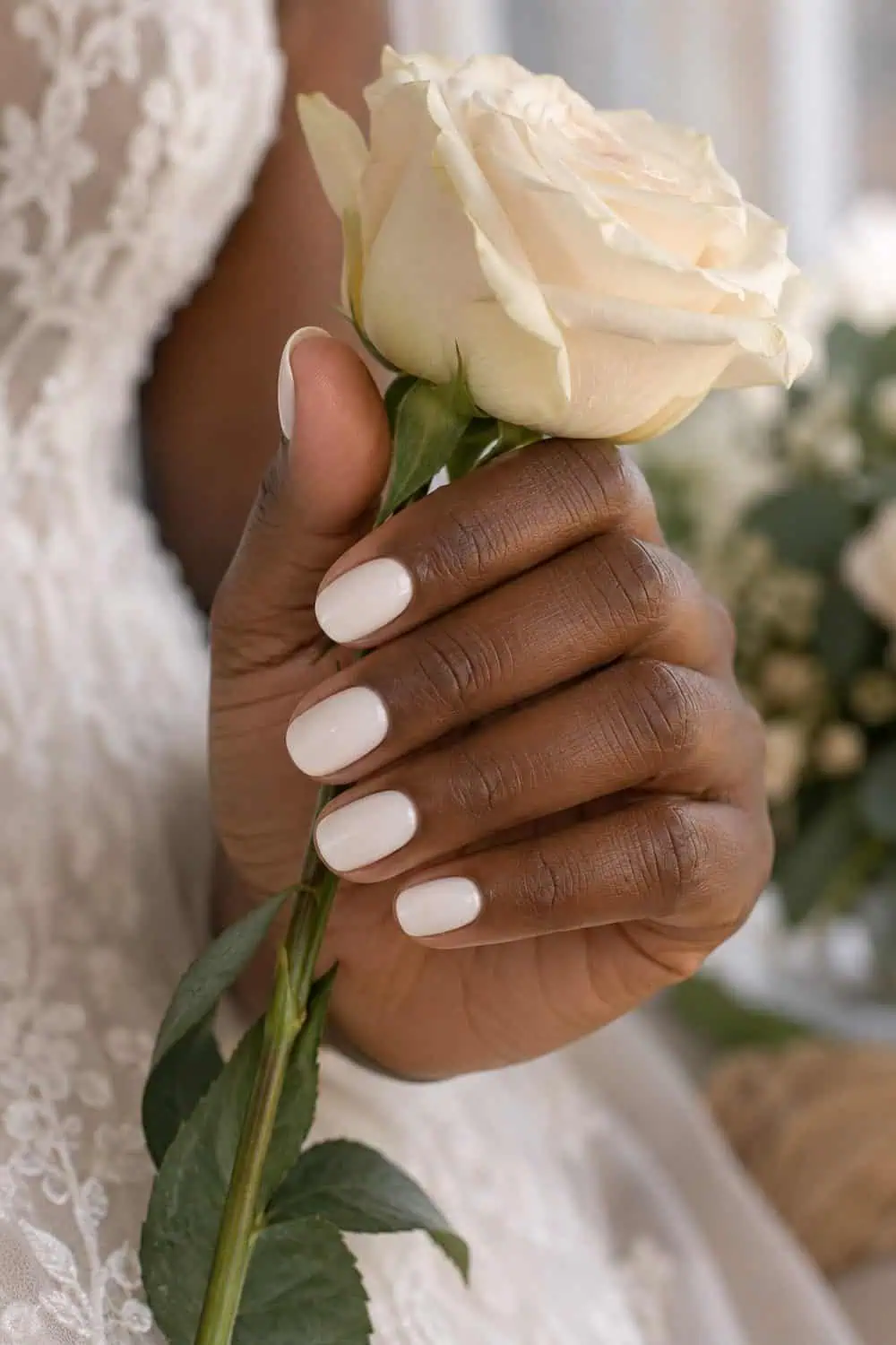 Bridal deep-brown hand with short oval nails painted smooth solid milky white, holding a cream rose