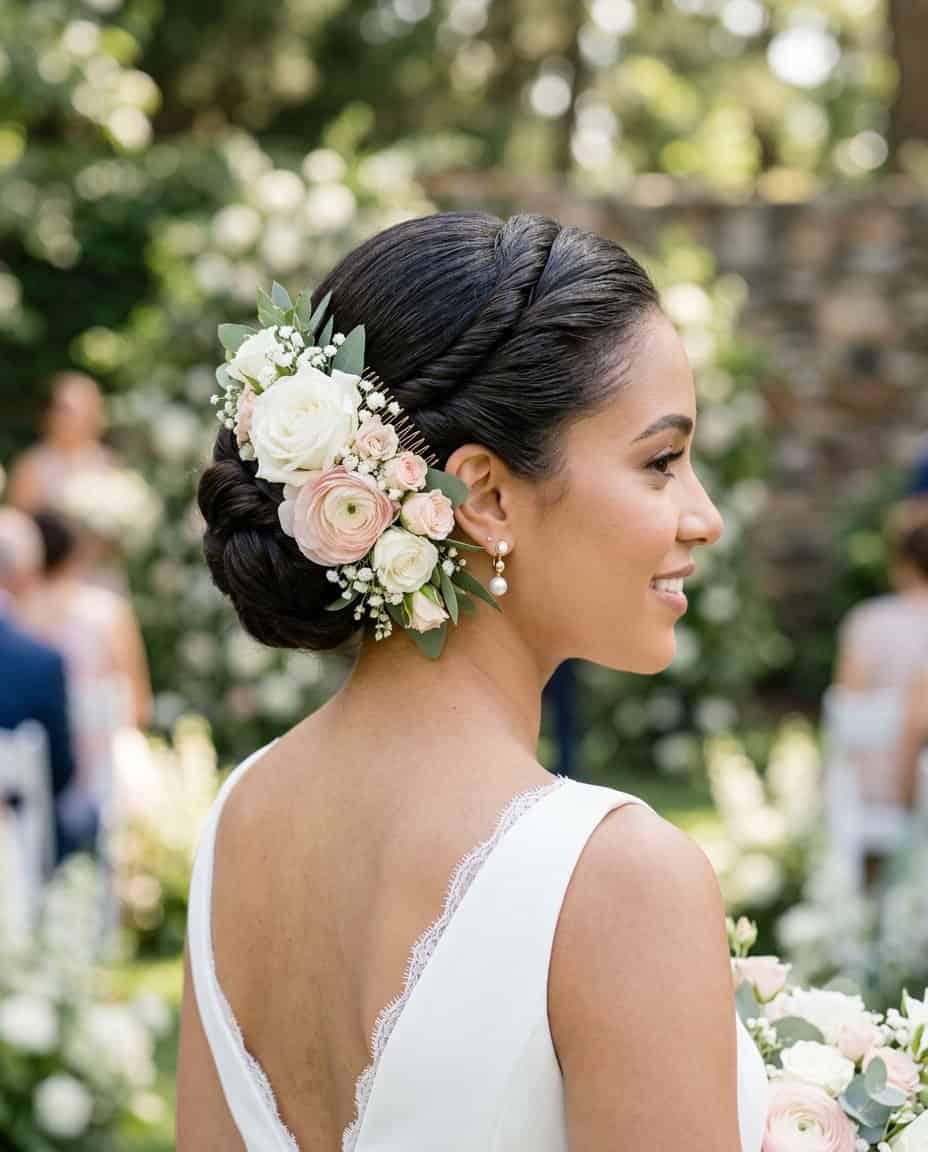 Close-up of a woman with flat twists shaped close to the scalp and gathered into a smooth bun at the nape, a bold floral comb placed just above it, wearing a white dress.