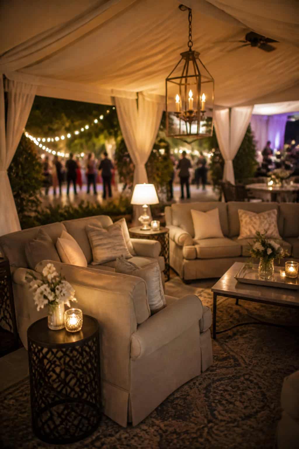 A covered lounge area with deep plush sofas, folded throw blankets, and a warm low-wattage lamp, positioned just off the edge of a reception tent