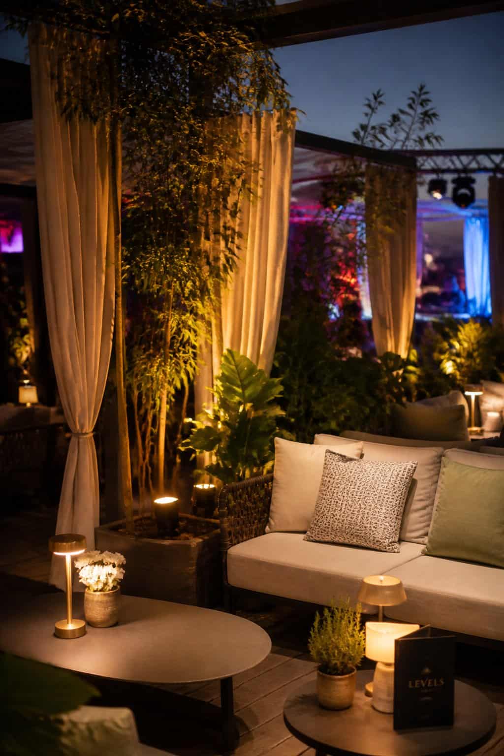 An outdoor pergola terrace with bistro tables, wicker loveseats draped in cream throws, warm string lights overhead, and tall potted ferns flanking linen panels between the terrace and the main ballroom entrance