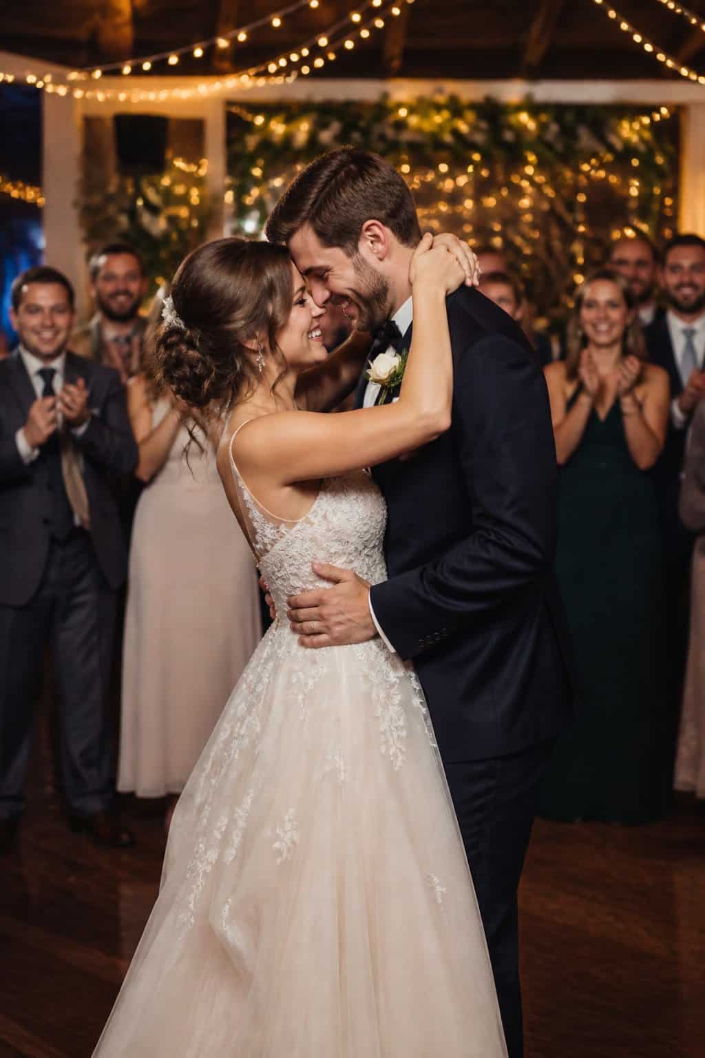 A newlywed couple smiling and dancing closely together at their wedding reception surrounded by applauding guests.