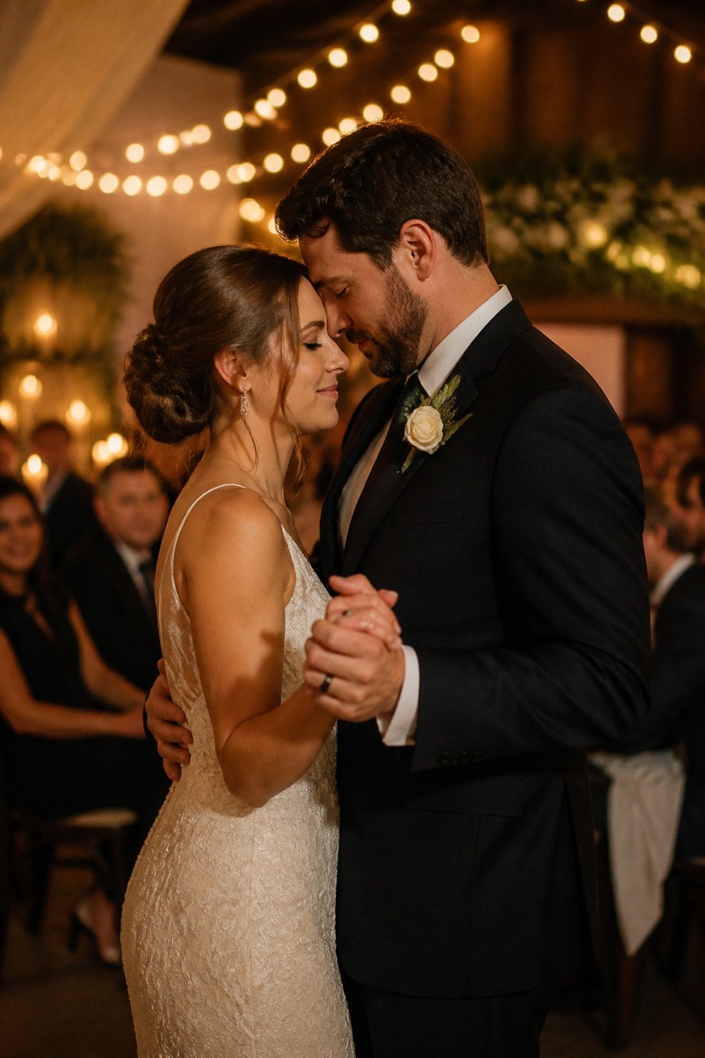 A couple sharing a tender second dance at a wedding reception with soft lighting and blurred guests around them.