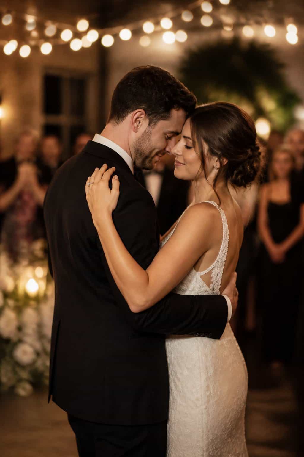 A couple embracing closely and dancing together at a wedding reception, surrounded by soft lights and blurred guests.