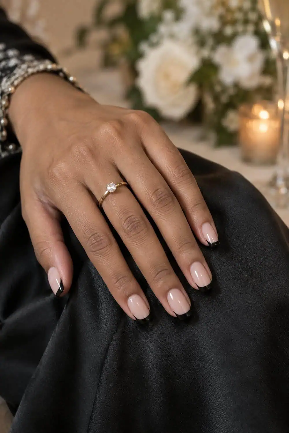 Wedding guest South Asian hand with medium soft-square nails, sheer beige base and slim black French tips, black satin fabric