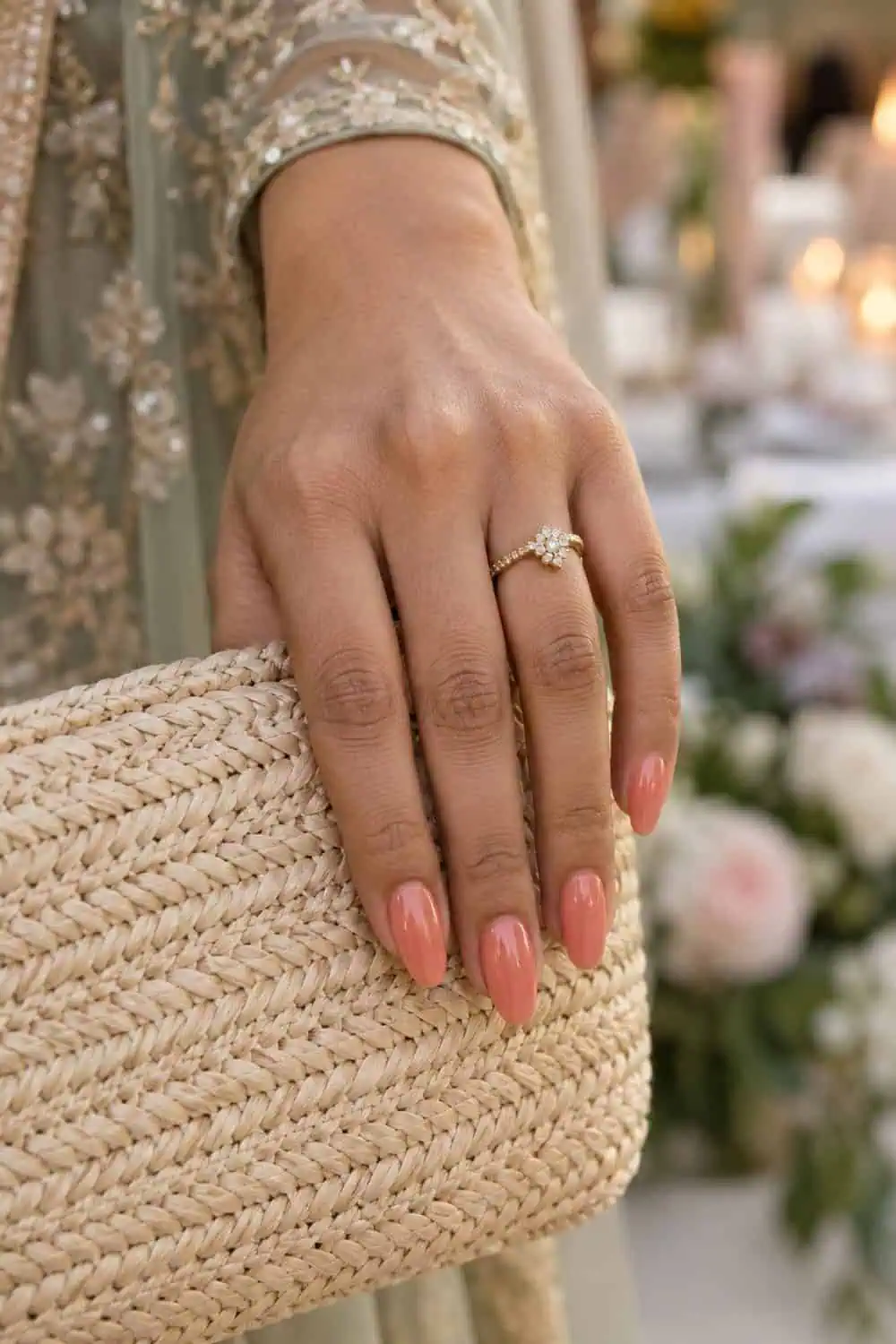 adult wedding guest's South Asian hand with medium almond nails painted translucent coral jelly polish, holding a woven neutral bag