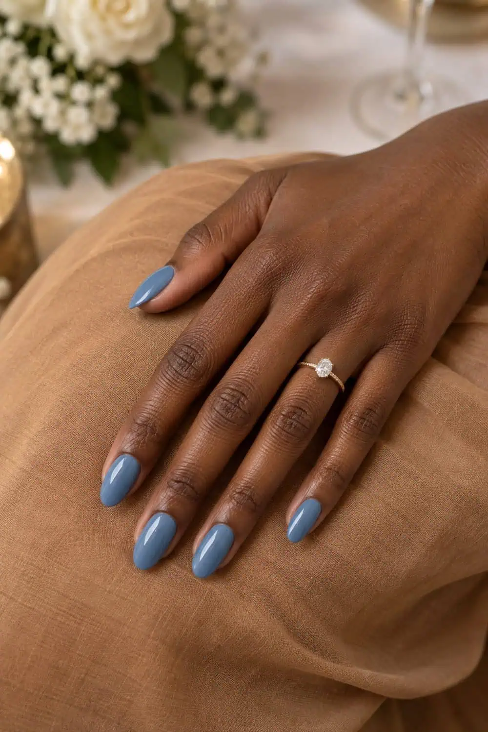 Wedding guest deep-brown hand with medium almond nails painted solid dusty blue, resting on camel-colored fabric