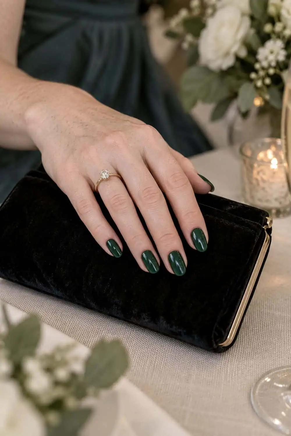 Wedding guest fair freckled hand with short almond nails painted solid deep emerald green, beside black velvet clutch