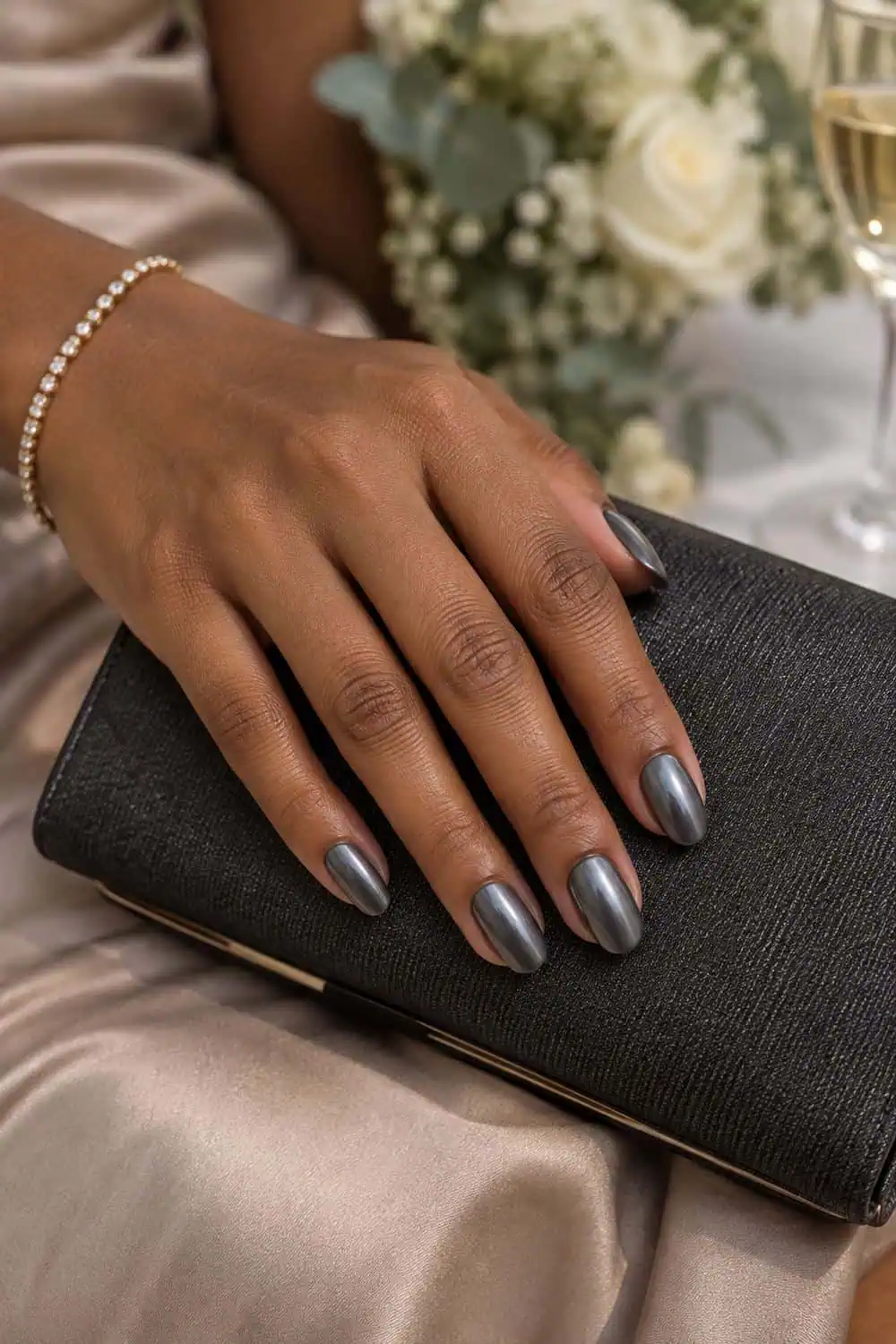 adult wedding guest's medium-brown hand with medium oval nails painted smooth muted pewter chrome, charcoal clutch nearby