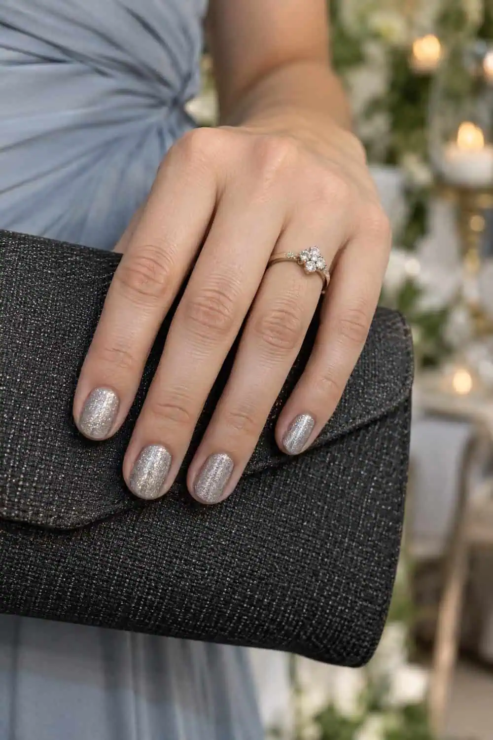 Wedding guest fair hand with short rounded nails coated in fine silver shimmer, holding a charcoal clutch