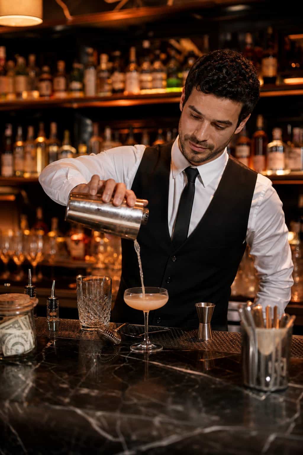 A bartender pouring a layered cocktail at a well-stocked bar with glassware and bar tools arranged on the counter.