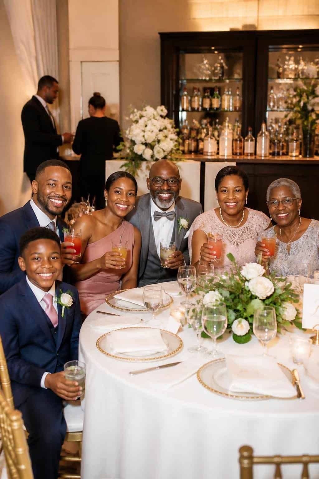 Family members seated together at a reception table, holding drinks with citrus garnishes and smiling, with floral centerpieces and string lights visible overhead.