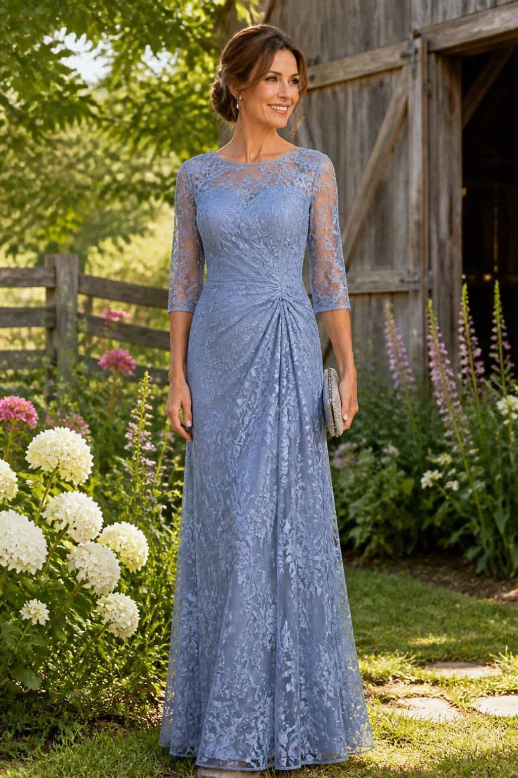 A woman in a full-length dusty blue lace gown with floral lace patterning over a chiffon skirt, standing near rustic wooden fencing with greenery behind her