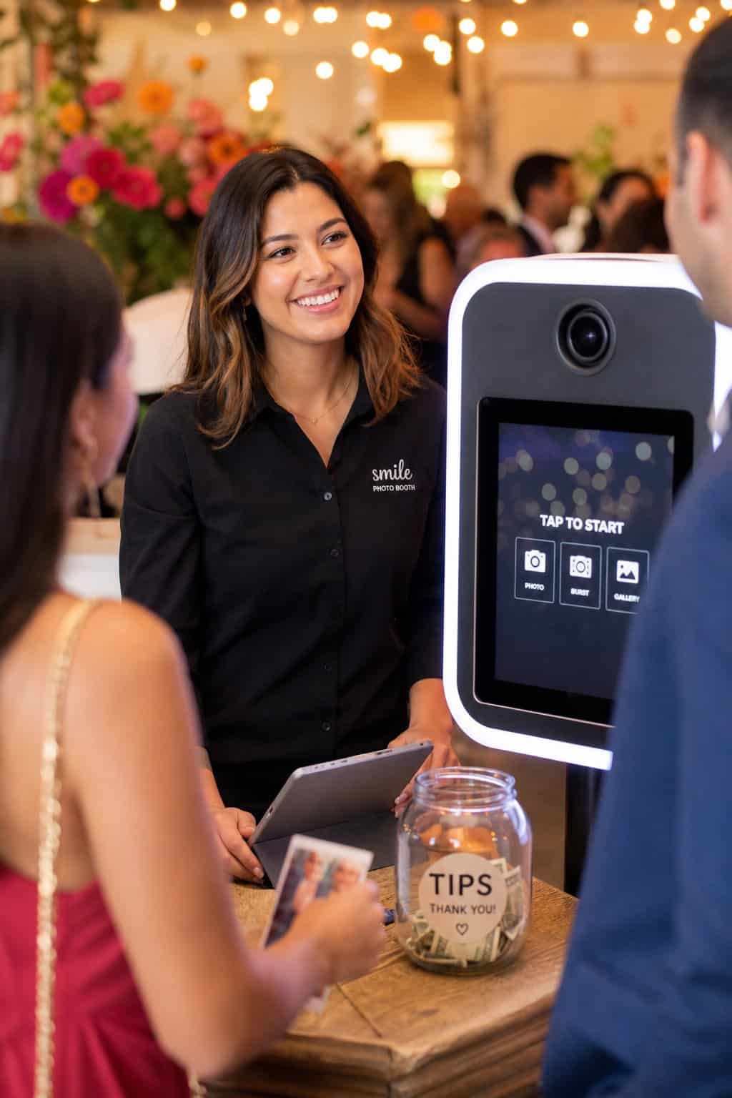 A smiling photo booth attendant standing behind a backdrop setup at an indoor reception, with a small tip jar visible on the prop table.