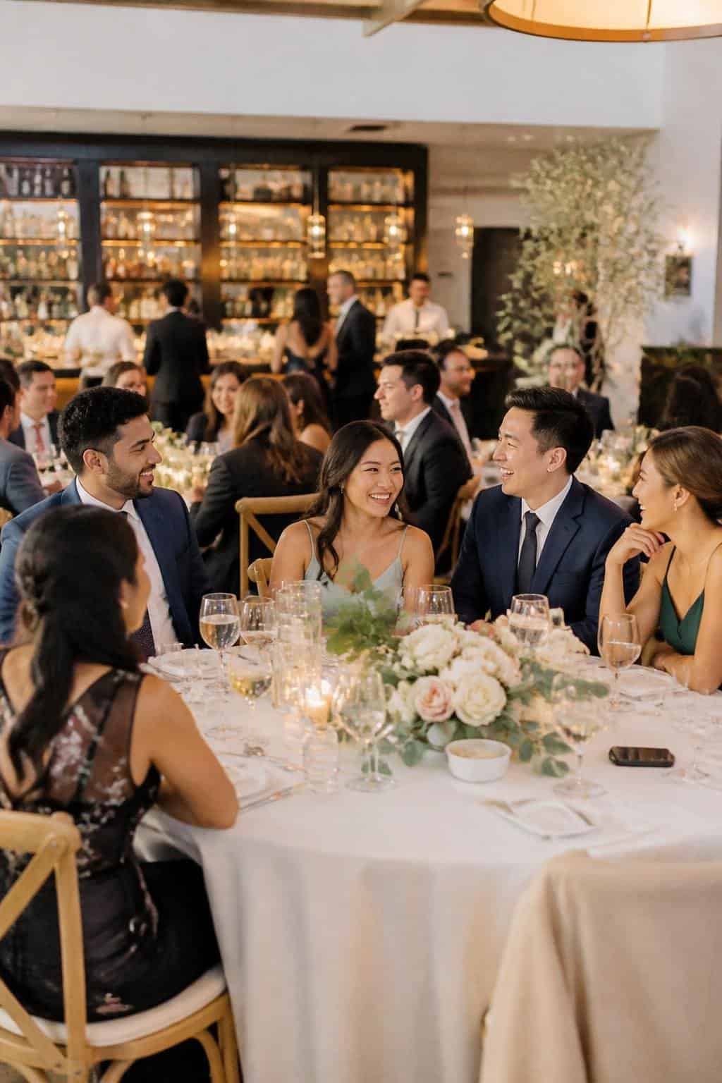 A mixed group of wedding guests including the groom's siblings and their partners seated together at a long table decorated with candles and greenery runners, laughing and talking.