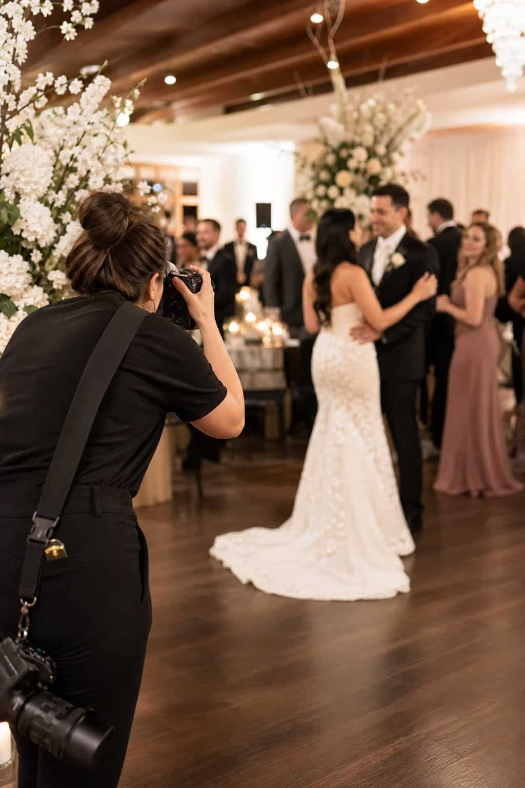 A photographer capturing candid moments at a wedding reception, with soft floral centerpieces and guests visible in the background.