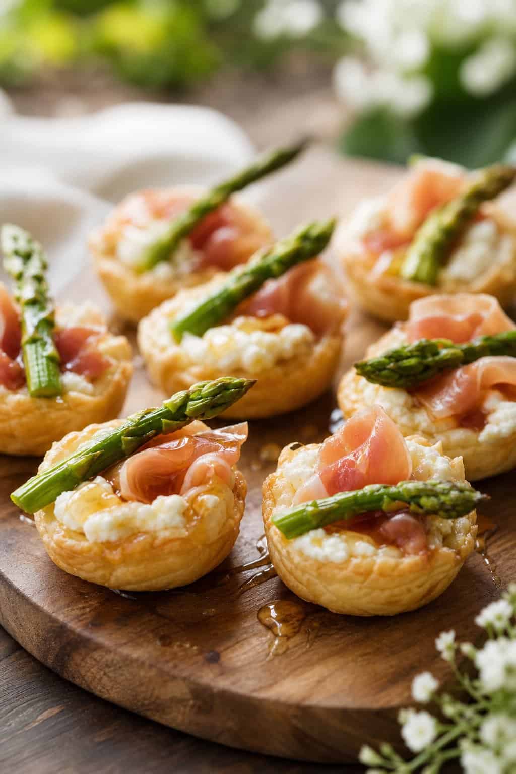 Golden puff pastry tartlets on a wooden board, each layered with white goat cheese spread, prosciutto-wrapped asparagus spears, and a glistening honey drizzle, shot in an outdoor setting.