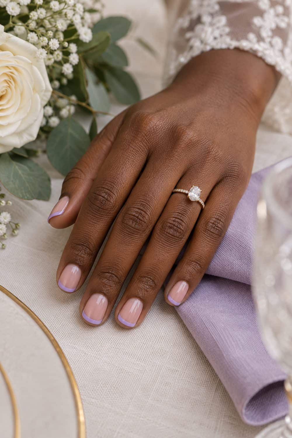 Bridal deep-brown hand with short square nails, sheer nude base and slim lavender French tips, lilac napkin nearby