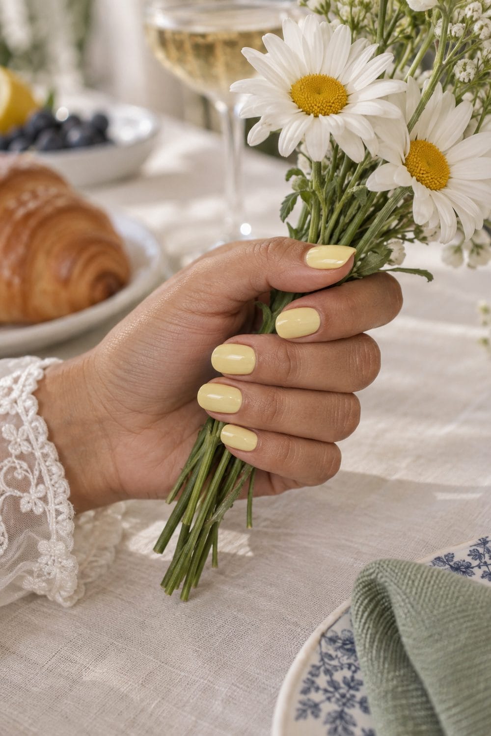 Bridal olive-toned hand with short oval nails painted solid soft lemon yellow, holding white daisies on a brunch table