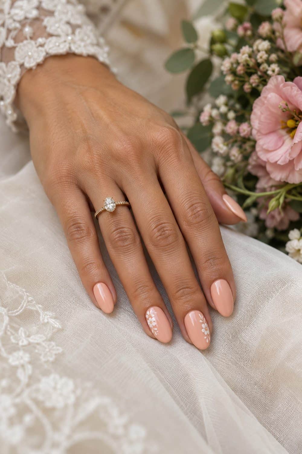adult bride's olive-toned hand with medium almond nails painted soft peach, tiny white floral corner details on two nails, pink flowers nearby