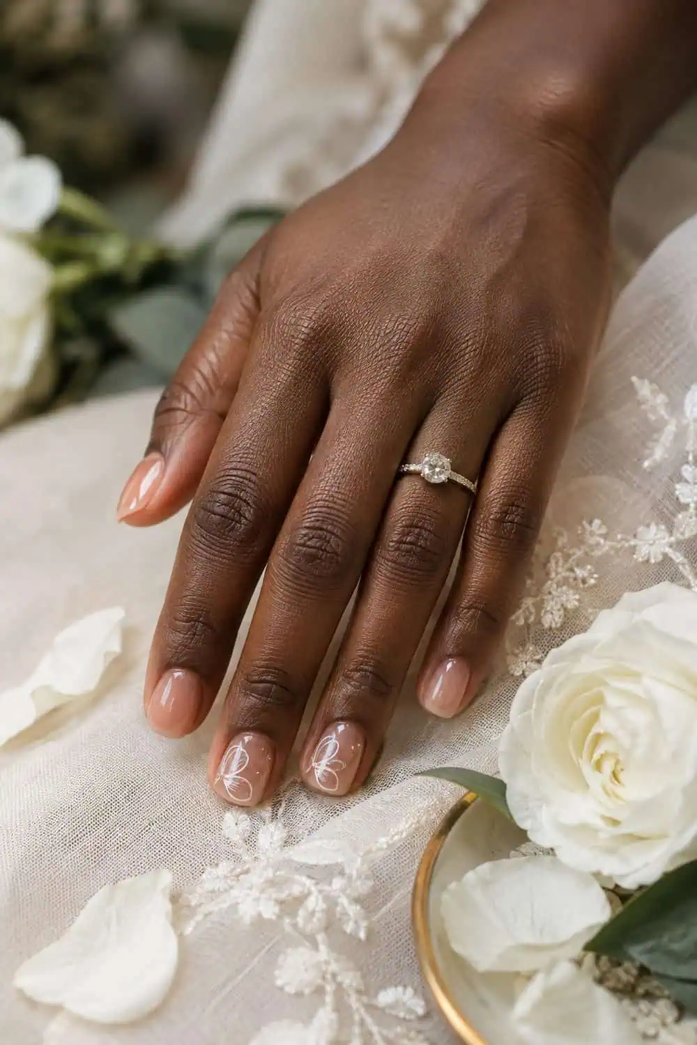 Bridal deep-brown hand with short squoval nails, clear gloss and delicate white petal outlines on two accent nails, real white petals nearby