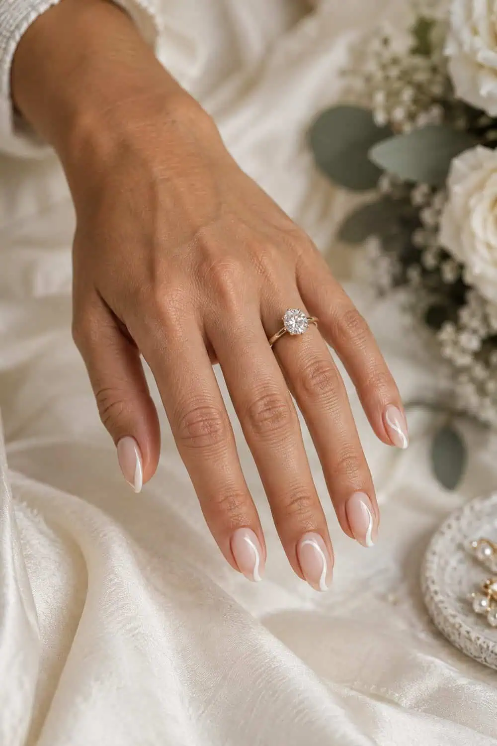 Bridal warm tan hand with medium oval nails, sheer nude base and slim curved ivory side-French detail along right edge, cream satin