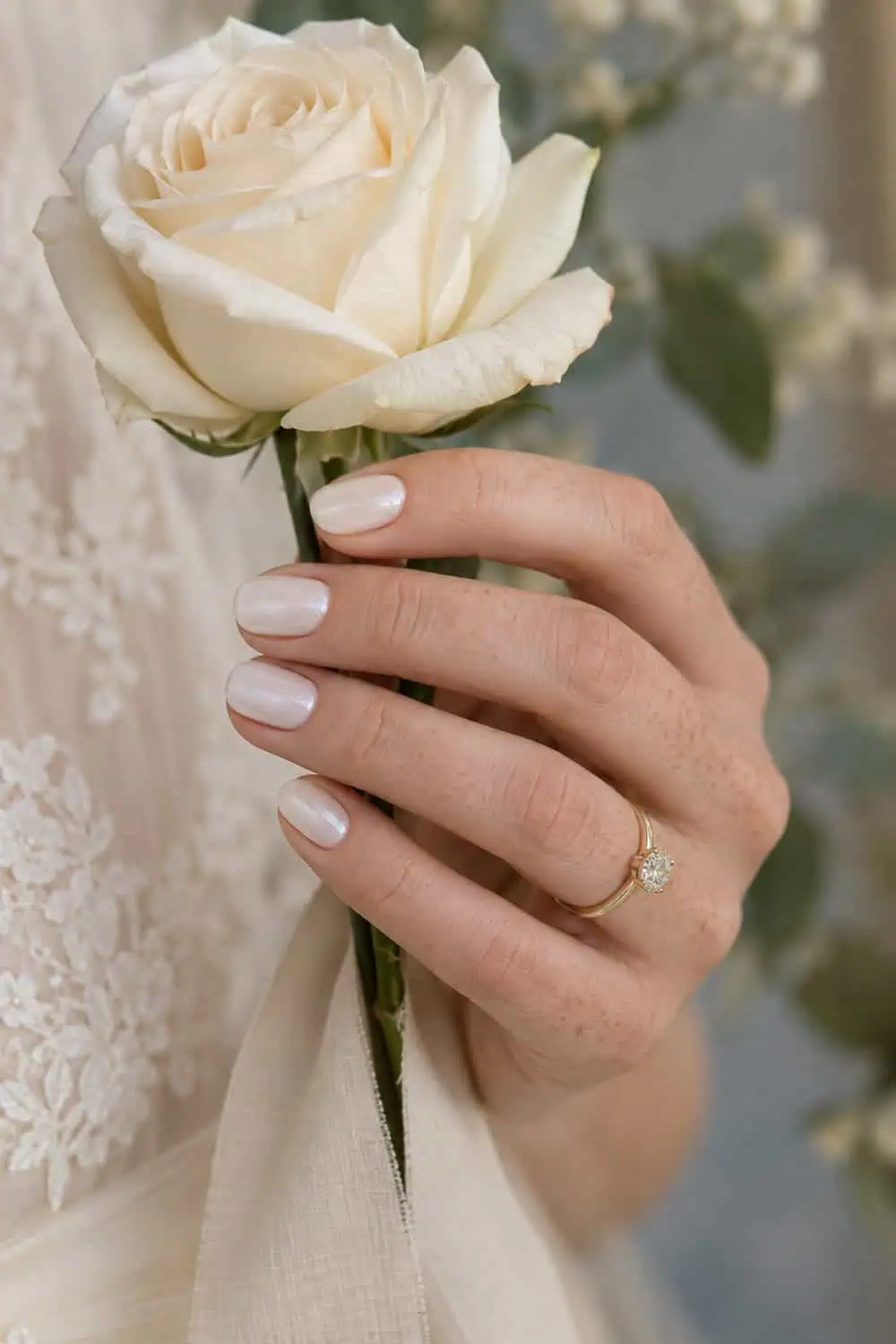 Bridal fair freckled hand with short oval nails painted smooth pearl white with fine even sheen, holding an ivory rose