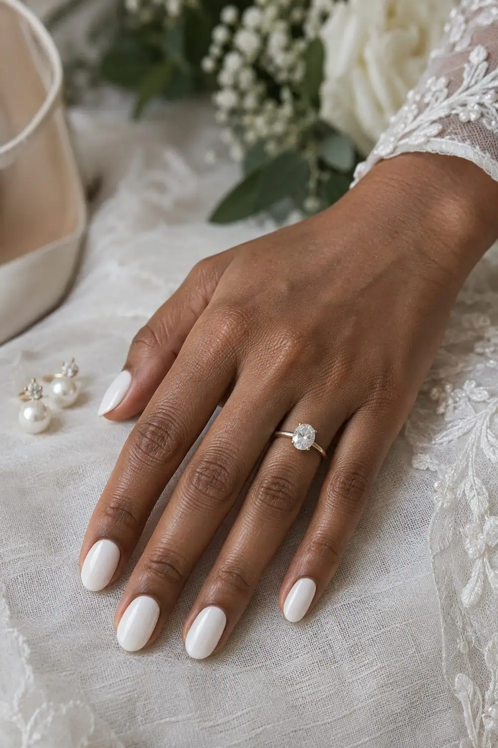 adult bride's medium-brown hand with medium oval nails painted smooth opaque porcelain white, pearl earrings nearby