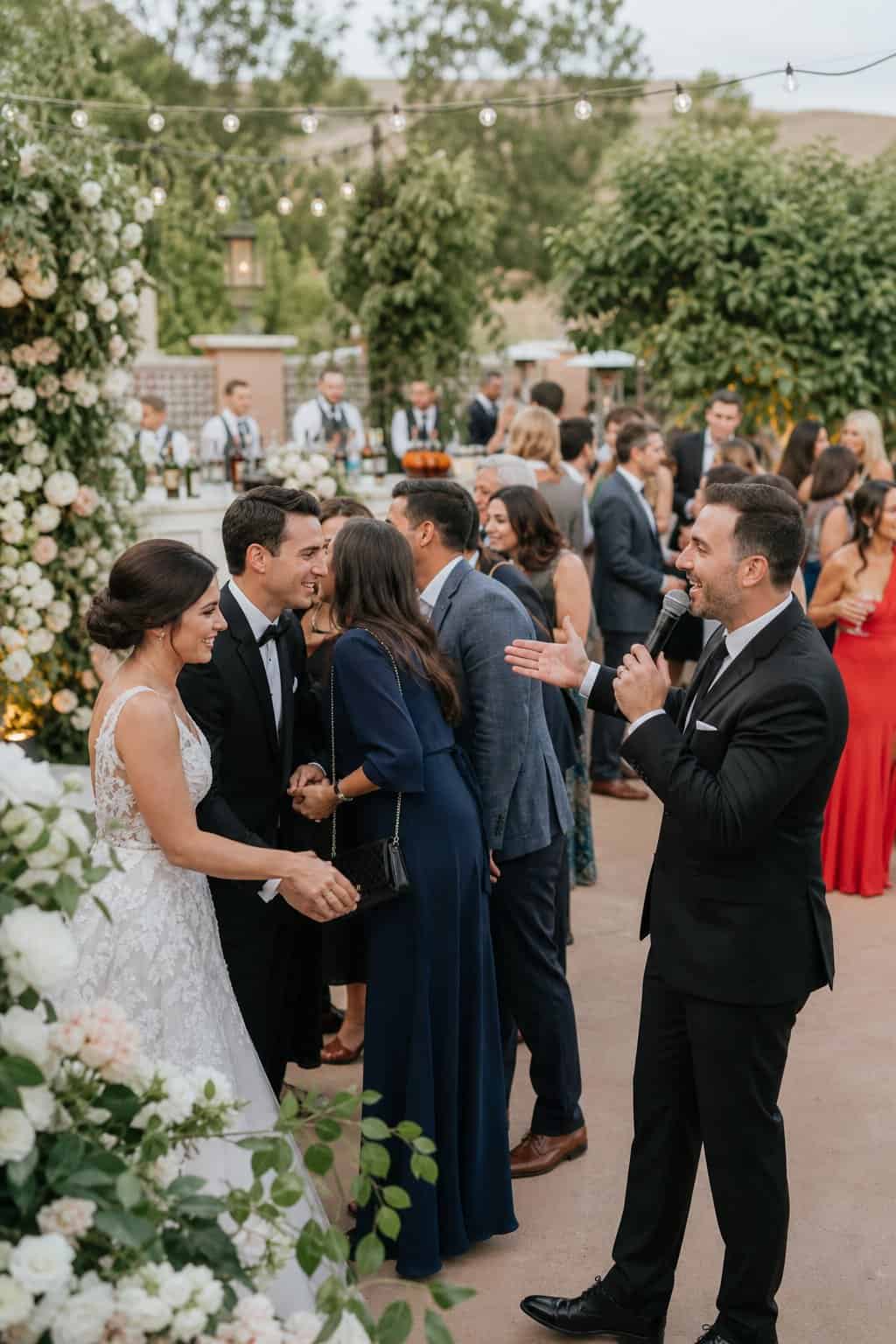 A DJ near an outdoor reception entrance calling groups of guests forward through a wedding receiving line while servers pass trays nearby.