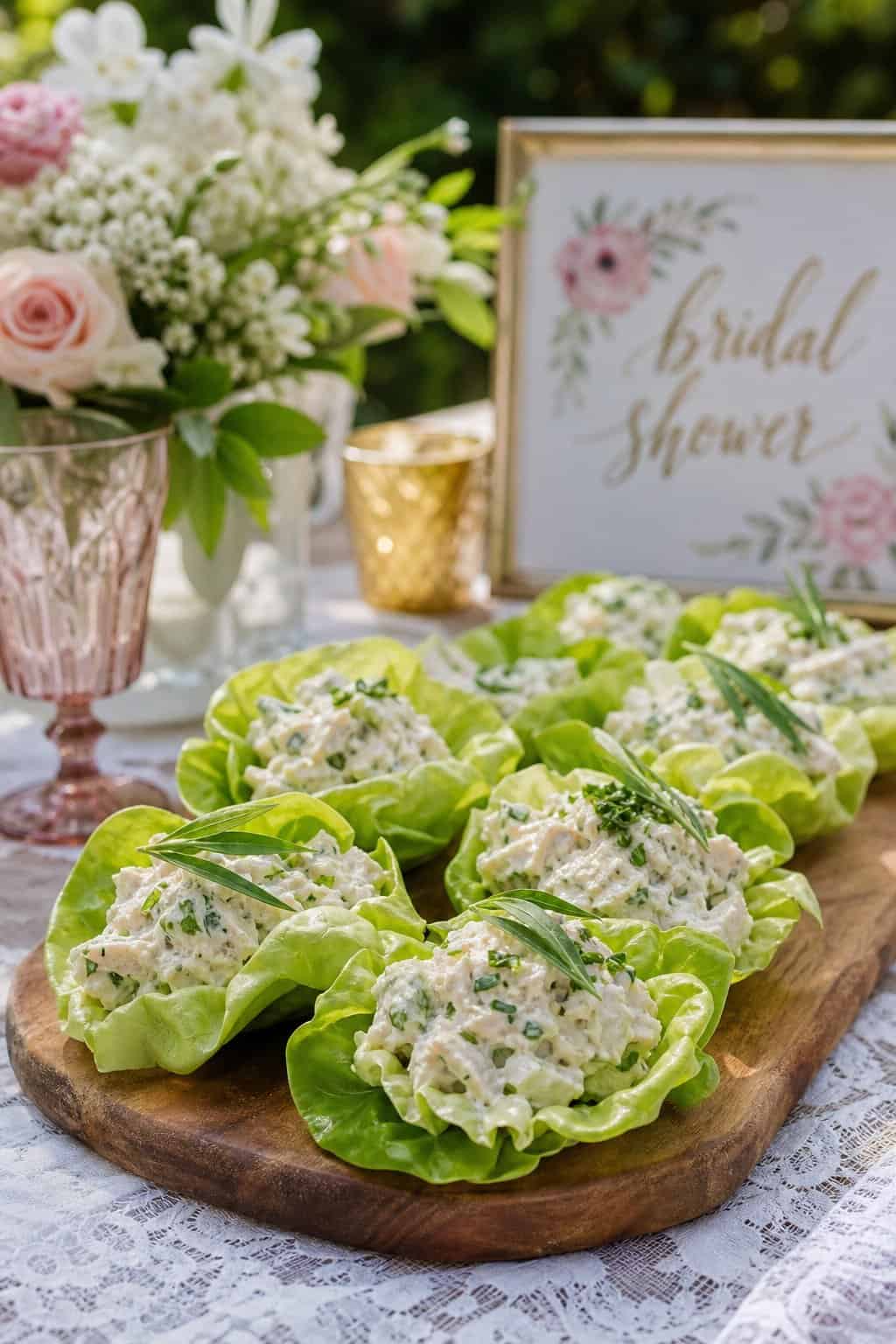 Butter lettuce cups cradling pale tarragon chicken salad on a wooden board, small flowers and glassware visible at the edges of the outdoor table.