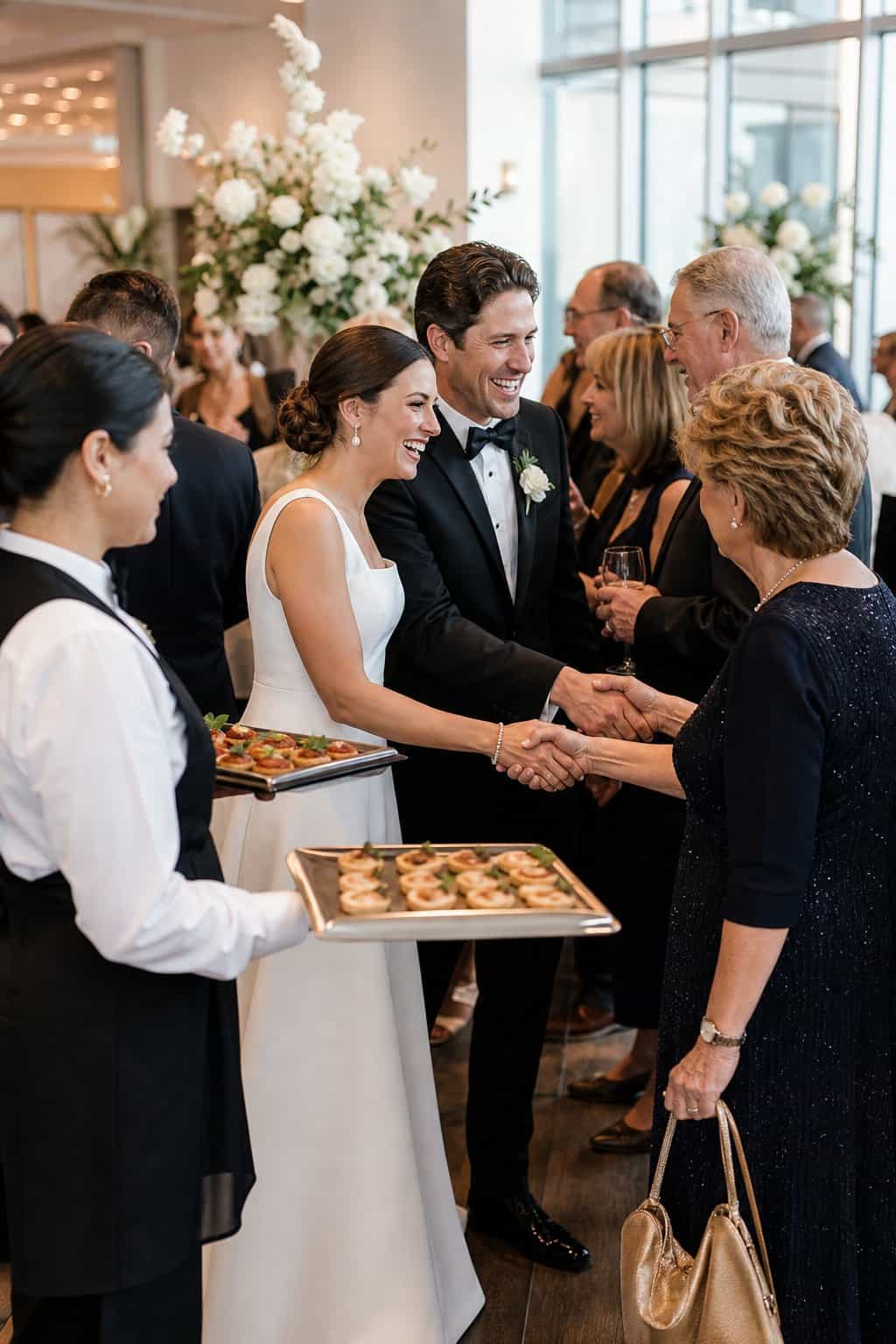 Servers in white shirts passing small hors d'oeuvres on trays to guests standing in a wedding receiving line in a well-lit reception hall.