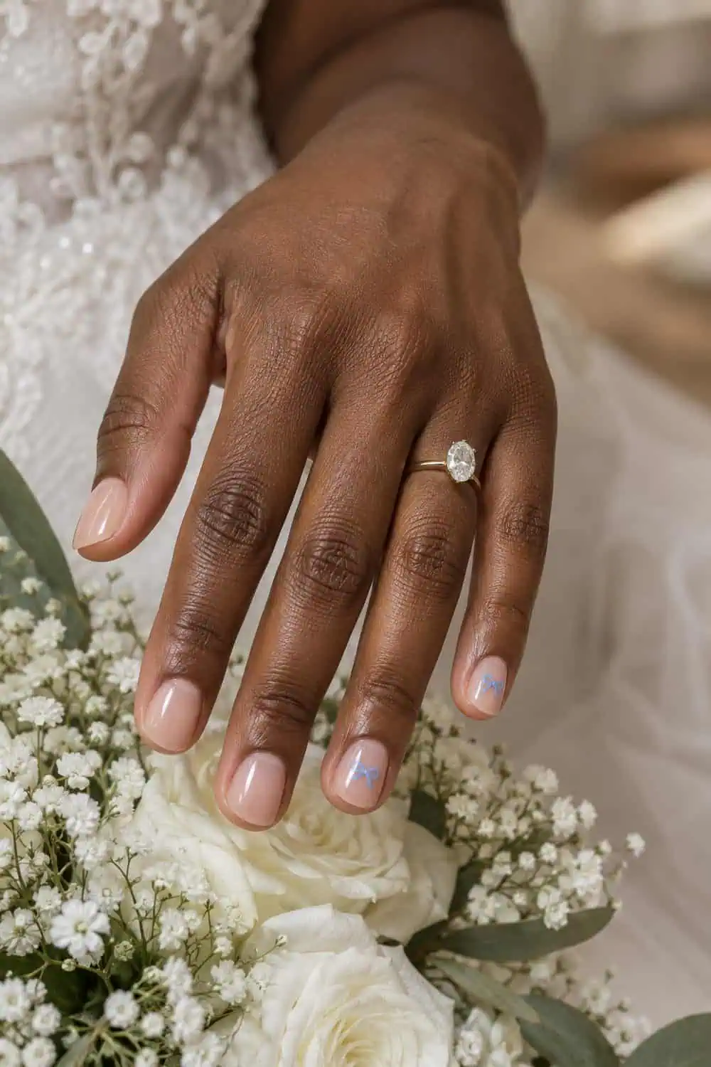 adult bride's medium-deep brown hand with short rounded nails under clear nude gloss, tiny flat baby-blue bows on two accents, white flowers