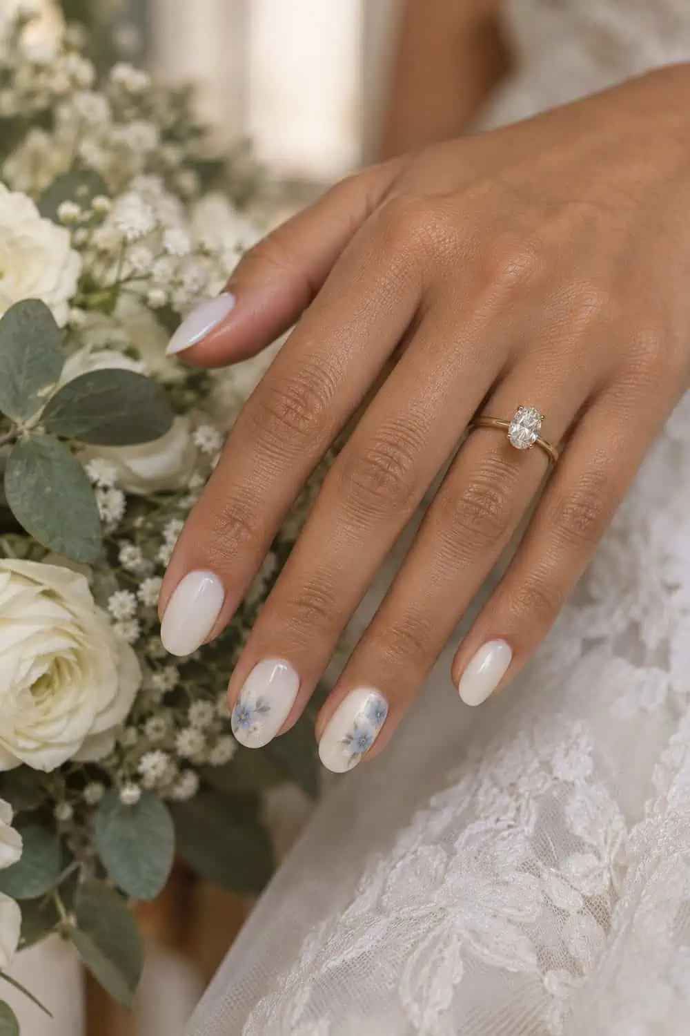 Bridal warm tan hand with medium oval nails, milky white base and small pale-blue floral corner clusters on two accents, white petals