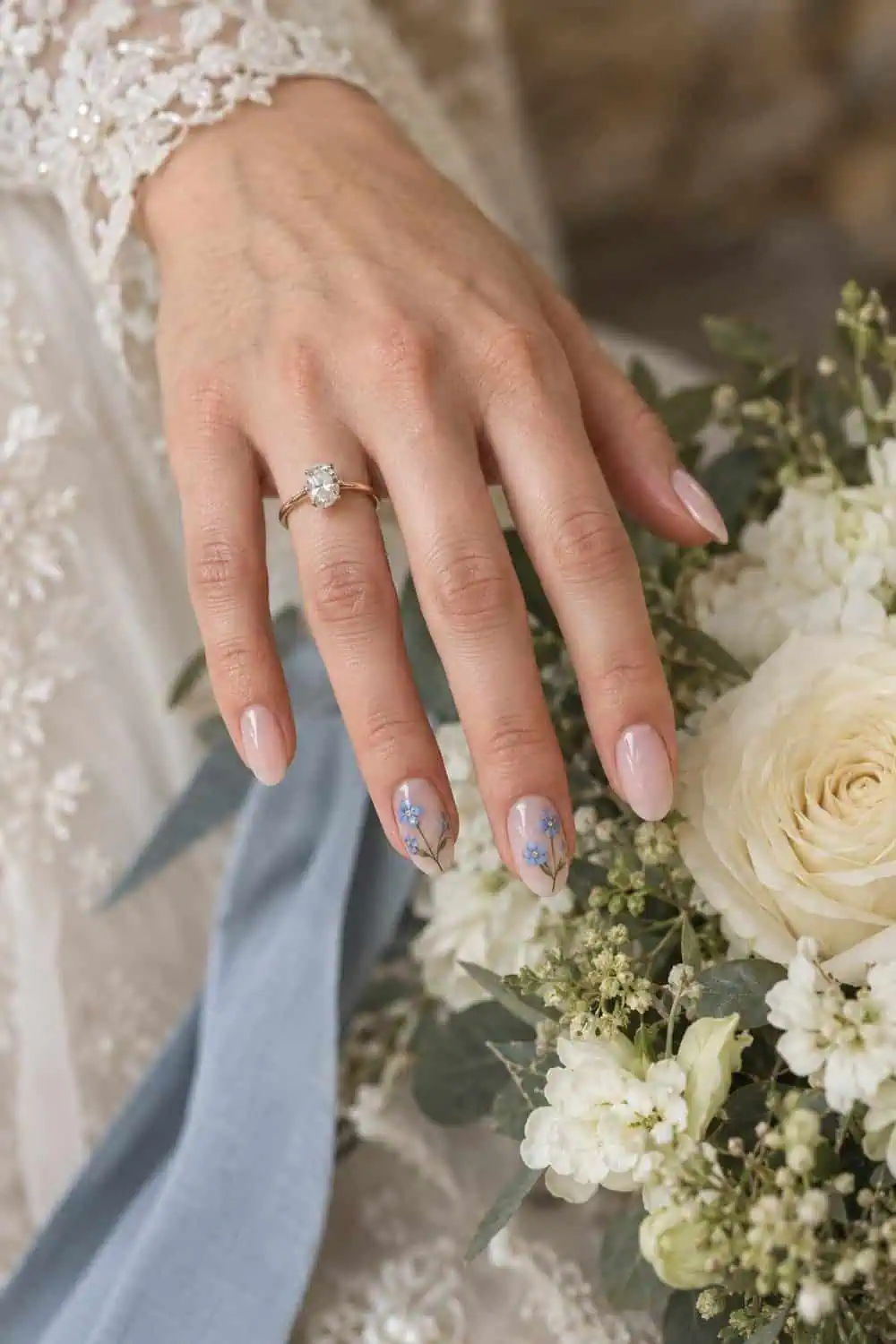 Bridal fair hand with medium oval nails, sheer pink base and tiny blue forget-me-not flowers on two accent nails, cream flowers nearby