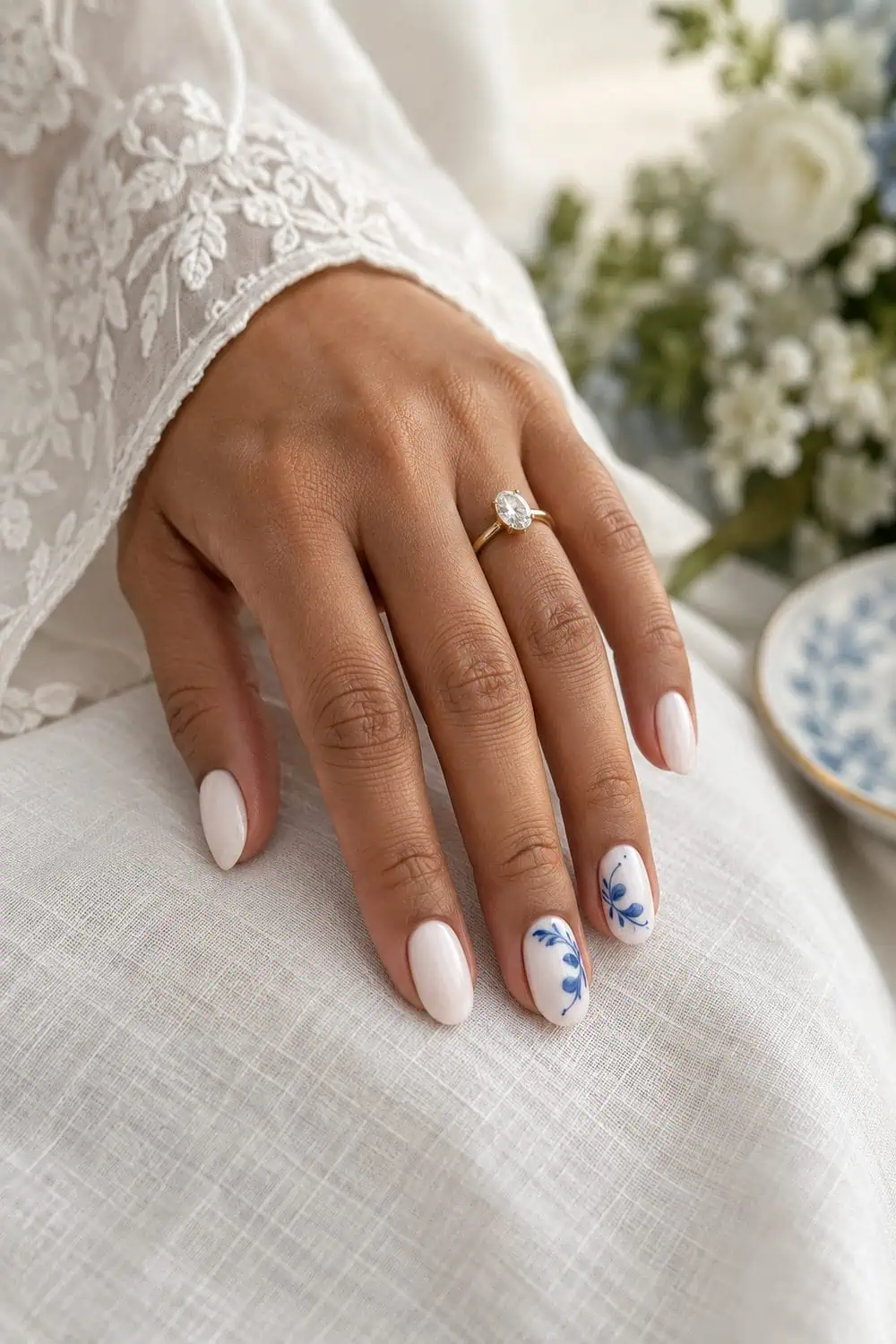 adult bride's light-brown hand with medium oval nails painted milky white, delicate cobalt-blue porcelain-inspired curves on two accent nails, white linen