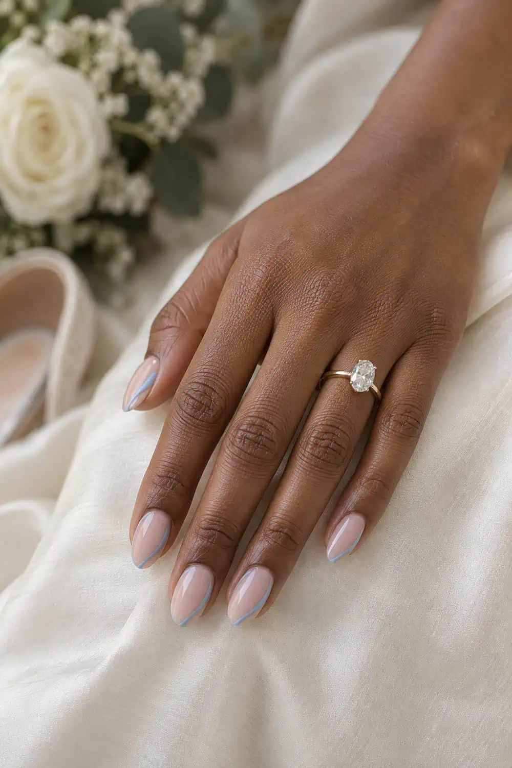 Bridal medium-deep brown hand with medium oval nails, sheer nude base and slim dusty-blue side-French curve, ivory silk