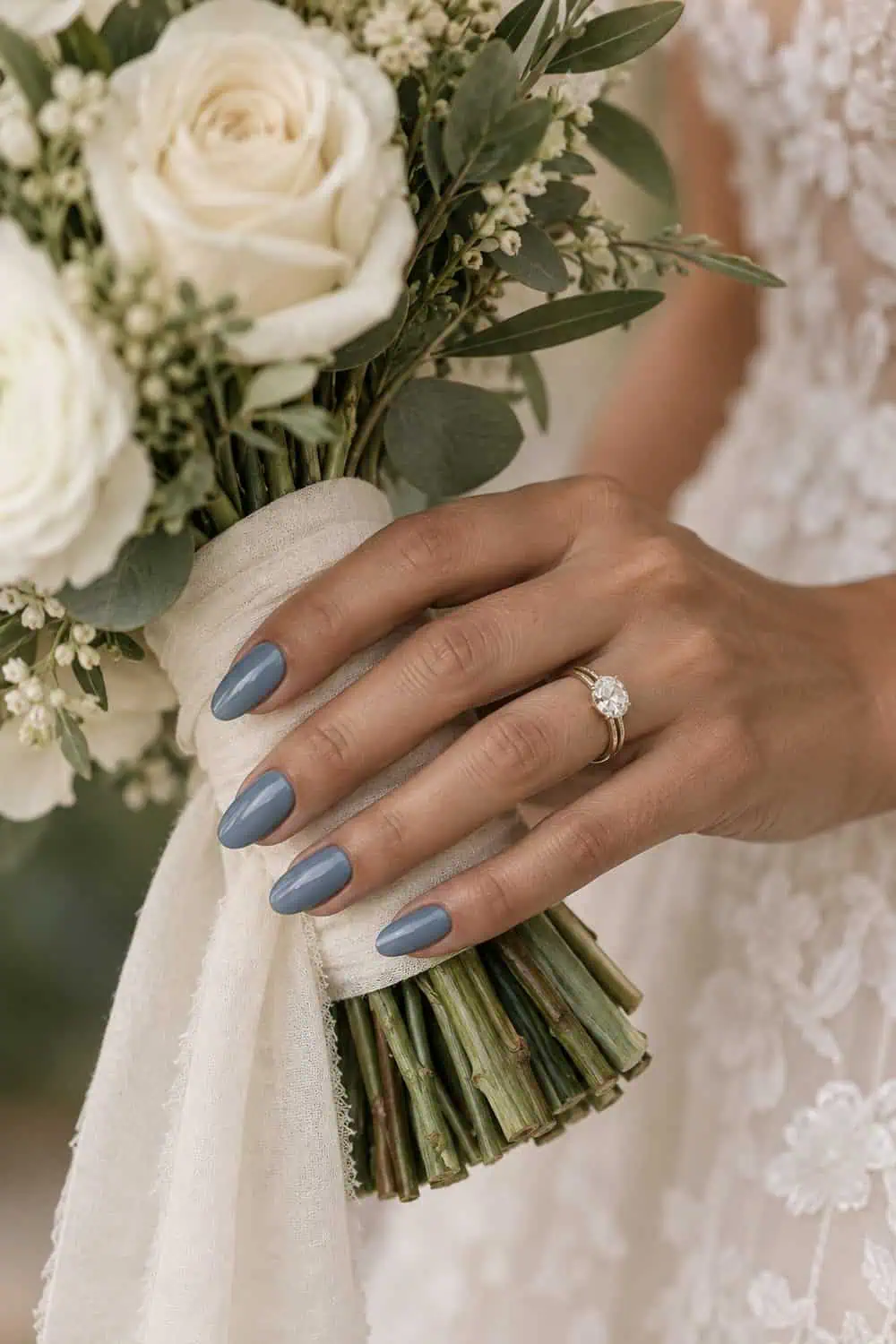 Bridal olive-toned hand with medium almond nails painted solid dusty blue, holding an ivory bouquet