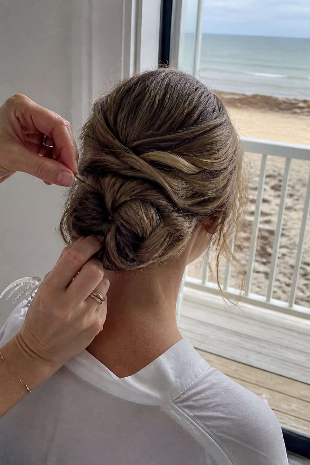 Hairstylist pinning a secure low braided bun in a room overlooking the beach