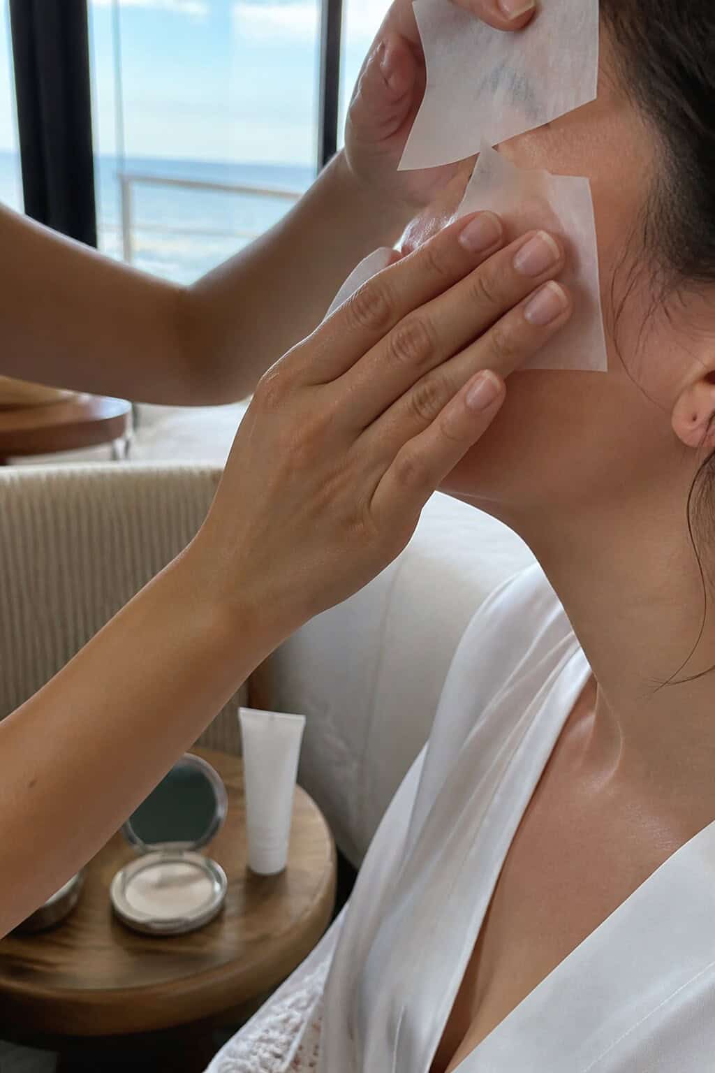 Makeup artist blotting a bride's skin beside a compact and sunscreen before a beach wedding