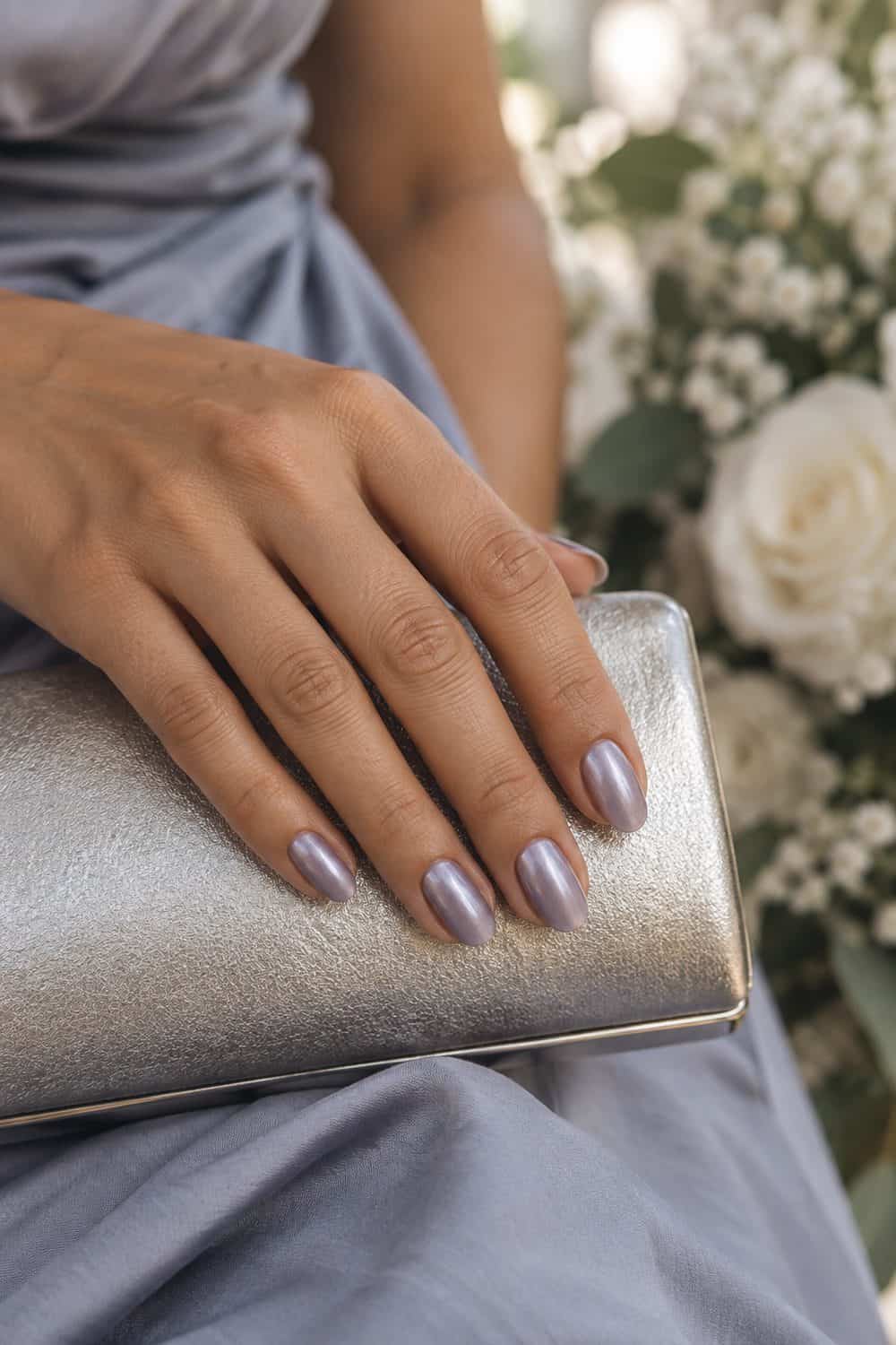 adult bridesmaid's olive-toned hand with medium oval nails painted dusty lavender with a subtle smooth chrome sheen, silver clutch nearby