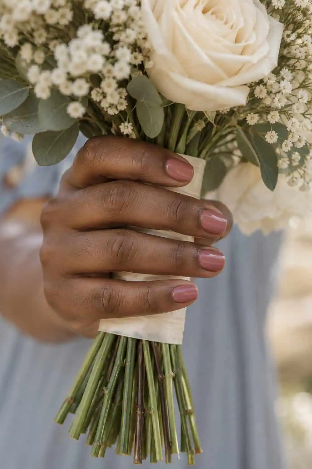 Bridesmaid deep-brown hand with short rounded nails painted solid dusty rose, holding a cream bridesmaid bouquet