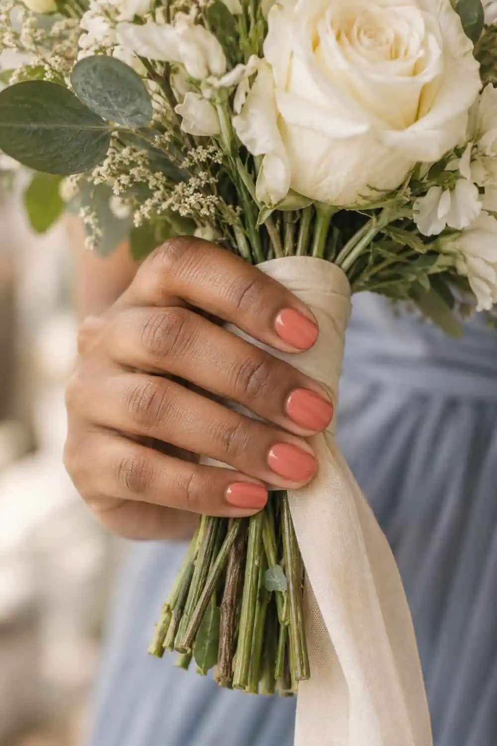 adult bridesmaid's medium-brown hand with short rounded nails painted solid muted coral, holding cream flowers
