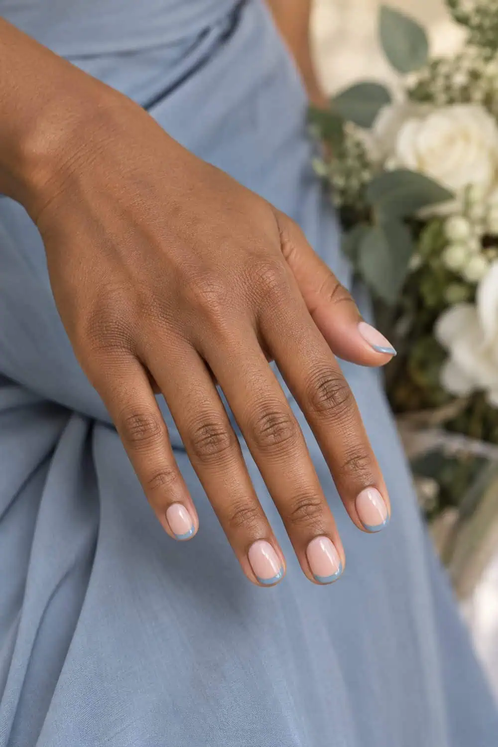 Bridesmaid medium-brown hand with short oval nails, sheer nude base and narrow dusty-blue French tips, dusty-blue dress fabric