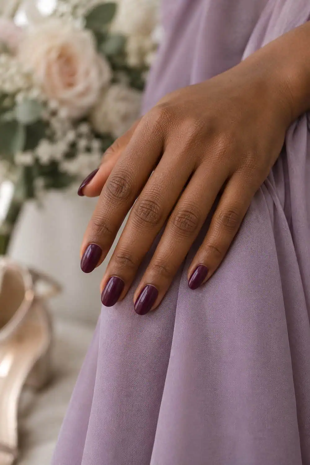Bridesmaid medium-deep brown hand with medium oval nails painted solid glossy plum, beside muted lilac fabric